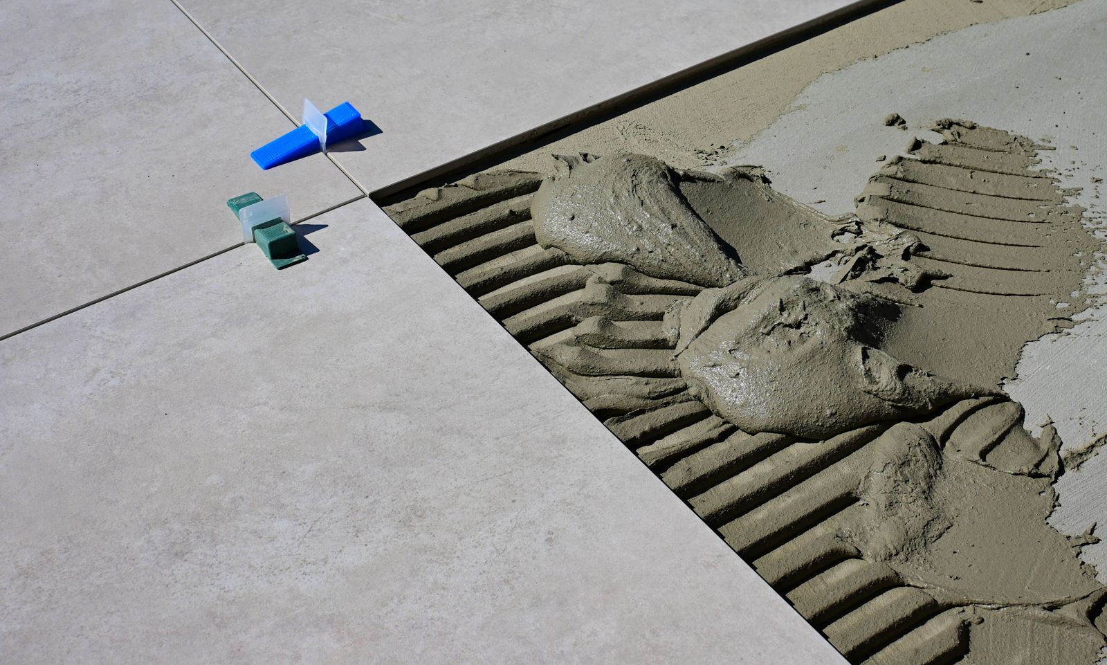 Large format floor tiles with leveling clips beside freshly combed tile adhesive